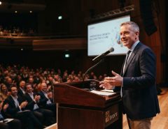 A speaker stands confidently at a podium on stage facing a warm audience.